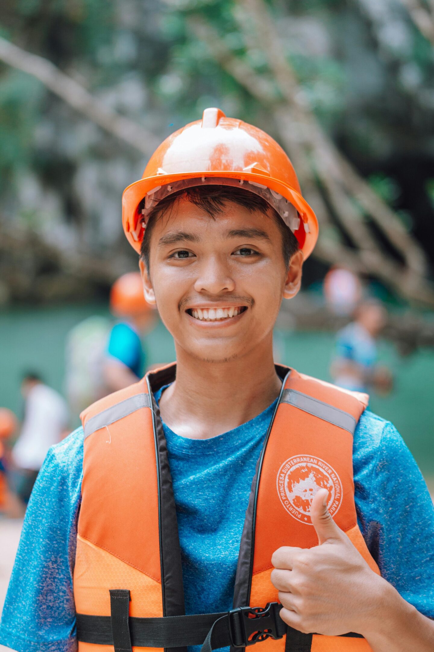 Smiling boy wearing an orange helmet and life jacket outdoors.